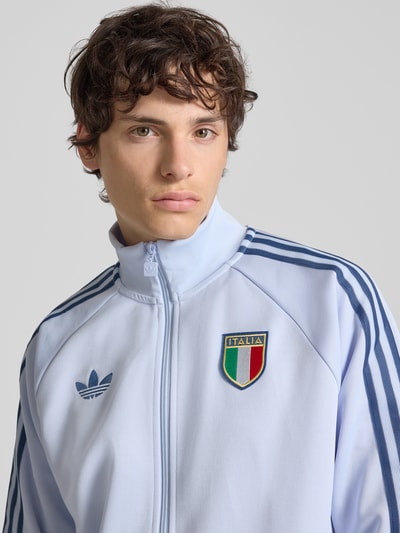Männermodel trägt Regular Fit Trainingsjacke mit Baumwoll-Anteil Modell 'AERBLU' von adidas Originals in Hellblau mit Italia-Plott, Logo-Stitching, Kontraststreifen und Stehkragen, Nahaufnahme Oberkörper.