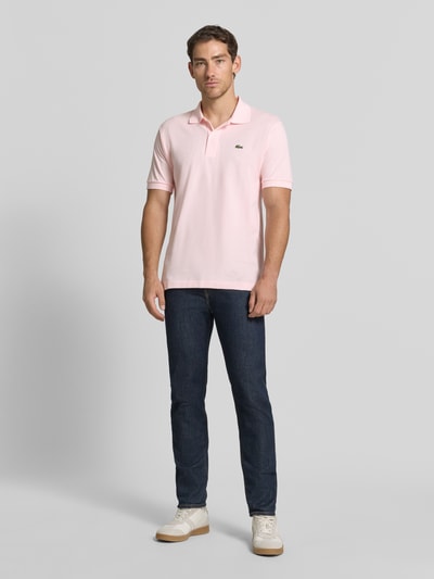 Männermodel trägt Regular Fit Poloshirt aus reiner Baumwolle von Lacoste in Hellrosa mit Kragen, kombiniert mit dunkler Jeans, Ganzkörperansicht von vorne, Herren Poloshirt von Lacoste.