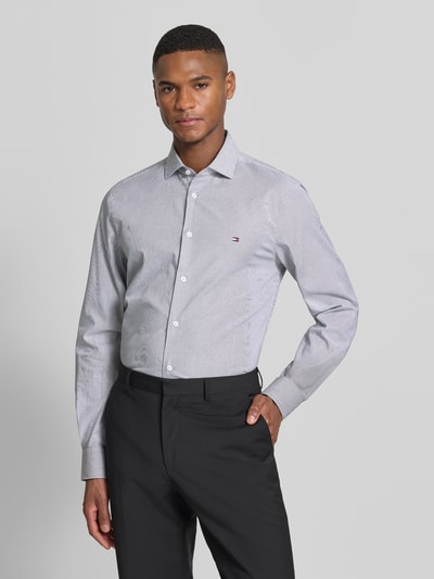 Männermodel trägt Slim Fit Businesshemd aus Baumwoll-Mix von Tommy Hilfiger, krój slim fit, wzór w paski, wyhaftowane logo, mieszanka bawełny, długi rękaw, mankiet zapinany na 1 guzik, kołnierzyk typu kent, listwa guzikowa na całej długości, ciemne spodnie, widoczne jasnoszare i białe paski, gładka tekstura, Ganzkörperansicht von vorne