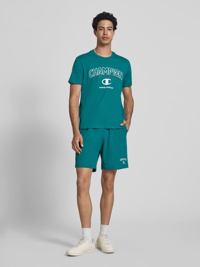 Männermodel trägt Sweatshorts mit Logo und elastischem Bund von CHAMPION, Regular Fit, Baumwoll-Mix, türkis, Ganzkörperansicht von vorne.