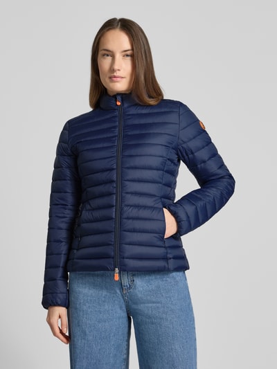 Frauenmodel trägt dunkelblaue Slim Fit Steppjacke mit gefüttertem Stehkragen Modell 'Carly' von SAVE THE DUCK, Stepp-Optik, Zweiwegereißverschluss, Eingrifftaschen mit Reißverschluss, Ganzkörperansicht von vorne.