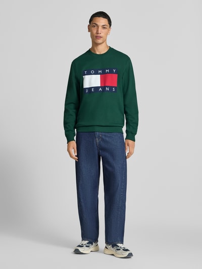 Männermodel trägt Regular Fit Sweatshirt aus reiner Baumwolle von Tommy Jeans in Dunkelgrün mit Logo-Print und blauer Jeans, Ganzkörperansicht von vorne. Herren Sweatshirt von Tommy Jeans.