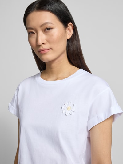 Modelka nosi T-Shirt mit floraler Applikation und fixiertem Ärmelaufschlag od Rich & Royal, damski t-shirt z bawełny ekologicznej, krój straight fit, okrągły dekolt, kwiatowa aplikacja, rękawy z zakładką, długość tyłu w rozmiarze S: 60 cm, zbliżenie na górną część ciała.