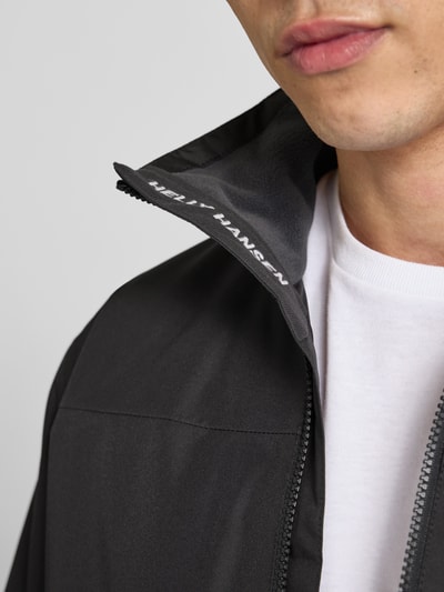 Mężczyzna nosi Midlayer Jacke mit Stehkragen Modell 'CREW' od Helly Hansen, czarny materiał, nadruk z logo, detal stójki, zamek błyskawiczny, zbliżenie na górną część kurtki