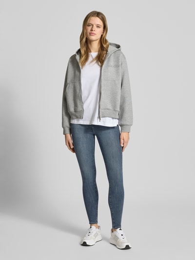 Frauenmodel trägt Skinny Fit Jeans aus Bio-Baumwoll-Mix Modell 'POWER' von Only in Blau, kombiniert mit weißem Shirt und grauer Kapuzenjacke, Denim-Optik, Ganzkörperansicht von vorne.