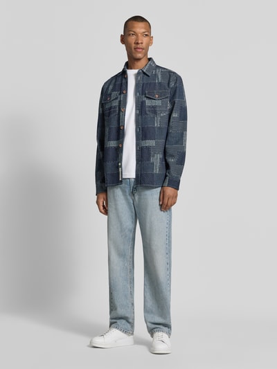 Männermodel trägt Relaxed Fit Hemdjacke in Denim-Optik von Scotch & Soda, Patchwork-Design, reine Baumwolle, Ganzkörperansicht von vorne.