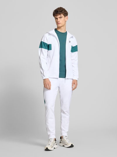 Mannenmodel draagt sweatpants met logo-stitching model 'CASPIO' van SERGIO TACCHINI, materiaalmix met stretch, nauwsluitend model, elastische band met trekkoord, zij-steekzakken, elastische boordjes, binnenbeenlengte maat M 78 cm, wit met groene accenten, gladde textuur, ganzkörperansicht van voren.