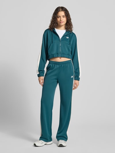 Frauenmodel trägt Sweatpants mit elastischem Bund und Label-Stitching von New Balance, Damen, Relaxed-Fit, Eingrifftaschen, petrol mit weißen Streifen, glatte Textur, Innenbeinlänge bei Größe S 80 cm, Ganzkörperansicht von vorne.
