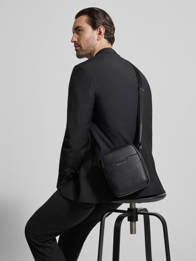 Männermodel trägt Handtasche mit verstellbarem Schulterriemen von ARMANI EXCHANGE, schwarze Struktur, Label-Detail, Ganzkörperansicht Rückansicht.