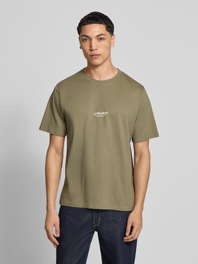Męski model nosi T-Shirt mit Logo und Rundhalsausschnitt od Jack & Jones, bawełna ekologiczna, krój regular fit, okrągły dekolt, logo na piersi, długość tyłu w rozmiarze M: 68 cm, oliwkowy, gładka faktura, widok całej sylwetki z przodu.