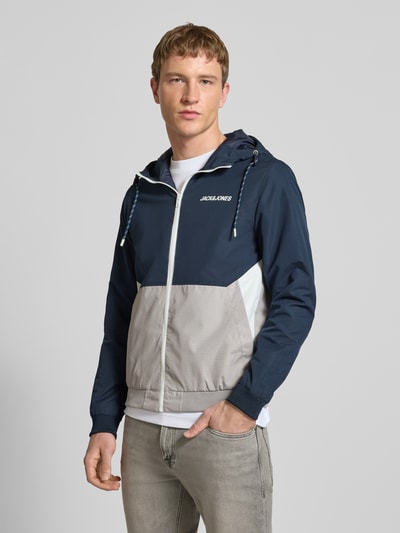 Mannelijk model draagt Jacke met capuchon en ritssluiting model 'RUSH' van JACK & JONES in regular fit, blauw en grijs, met logoprint, steekzakken, ruglengte bij maat M 68 cm, schouderbreedte 46 cm, borstwijdte 55 cm, mouwlengte 68 cm, voorkant bovenlichaam close-up.
