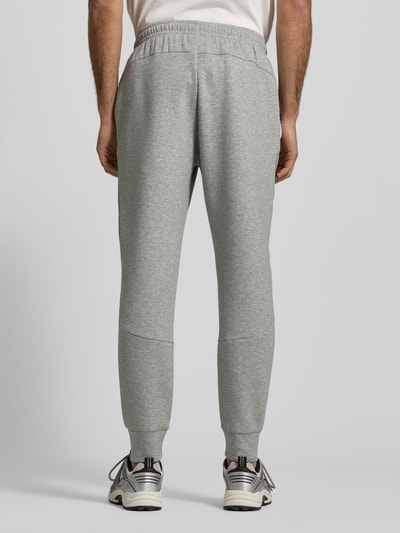 Männermodel trägt Regular Fit Sweatpants mit Logo-Print Modell 'Unstoppable' von Under Armour in Hellgrau mit elastischem Bund und Beinabschlüssen, Ganzkörperansicht Rückansicht. Elastischer Material-Mix mit Baumwoll-Anteil, Beintasche mit Reißverschluss.