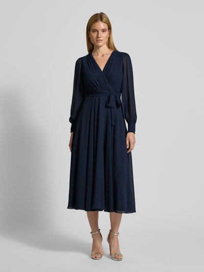 Frauenmodel trägt Regular Fit Midikleid mit Taillenband von Christian Berg Cocktail aus navyfarbenem, semitransparentem Chiffon mit blickdichtem Unterrock, V-Ausschnitt und Wickel-Optik, Falten-Textur, langärmelig; Ganzkörperansicht von vorne.
