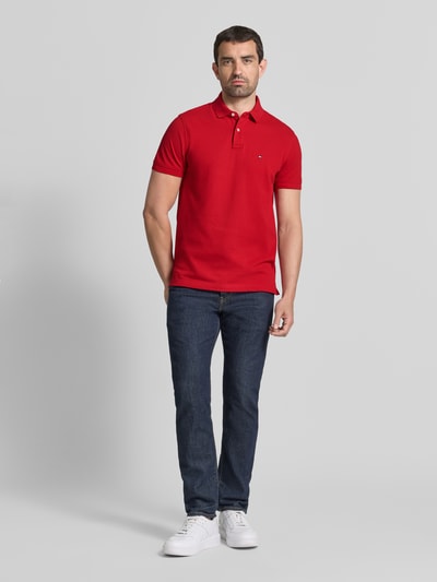 Männermodel trägt Regular Fit Poloshirt aus Baumwoll-Mix von Tommy Hilfiger in Rot, kombiniert mit dunkler Jeans, Ganzkörperansicht von vorne.