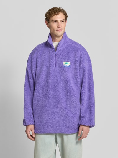 Männermodel trägt Oversized Pullover mit Stehkragen Modell 'HOKTOWN' von American Vintage in Lila, Kunstfell, Logo-Patch, Ganzkörperansicht von vorne. Rückenlänge 85 cm, Ärmellänge 60 cm, elastische Abschlüsse.