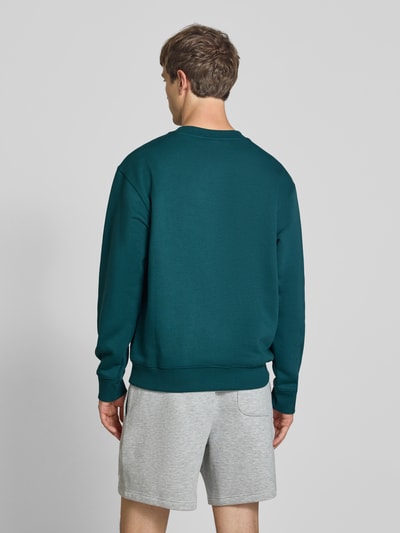 Männermodel trägt Sweatshirt mit Rundhalsausschnitt und Label-Stitching von New Balance in Dunkelgrün, Baumwoll-Mix, Ganzkörperansicht Rückansicht.