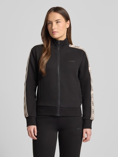 Frauenmodel trägt Regular Fit Sweatjacke mit Stehkragen Modell 'Britney' von Guess Activewear, Schwarz, Logo-Detail, Ganzkörperansicht von vorne.