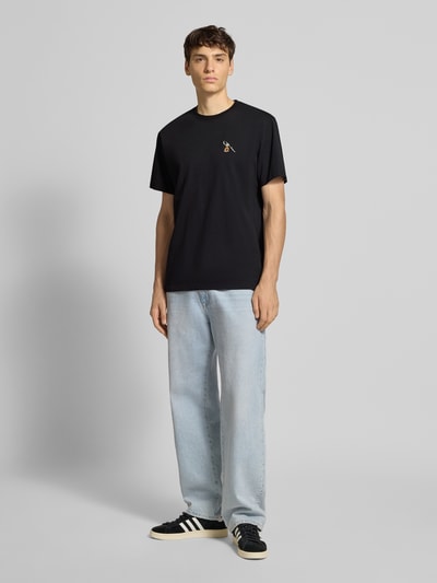 Męski model nosi T-Shirt mit Motiv-Stitching od Dedicated, czarna bawełna, krój regular fit, haft z przodu, widok całej sylwetki z przodu.