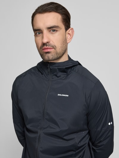 Männermodel trägt Windbreaker mit Kapuze und Logo von Salomon aus Material-Mix mit Stretch-Anteil, Regular-Fit, Nahaufnahme Oberkörper.