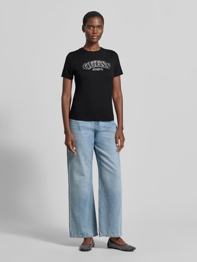 Frauenmodel trägt T-Shirt aus Viskose-Elasthan-Mix mit Logo von Guess Activewear in Schwarz, kombiniert mit Jeans, Ganzkörperansicht von vorne.