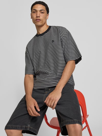 Männermodel trägt Carhartt Work In Progress Herren T-Shirt mit Logo-Stitching Modell 'Dupree' aus Reine Baumwolle, Boxy-Fit, Rundhalsausschnitt, horizontale Streifen in Schwarz und Weiß, strukturierte Textur, Rückenlänge 70 cm bei Größe M, Ganzkörperansicht von vorne.
