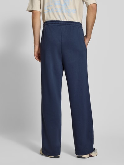 Männermodel trägt blaue Sweatpants mit Logo-Stitching und Eingrifftaschen von Karl Kani, Baumwoll-Mix, Wide Fit, Ganzkörperansicht Rückansicht.