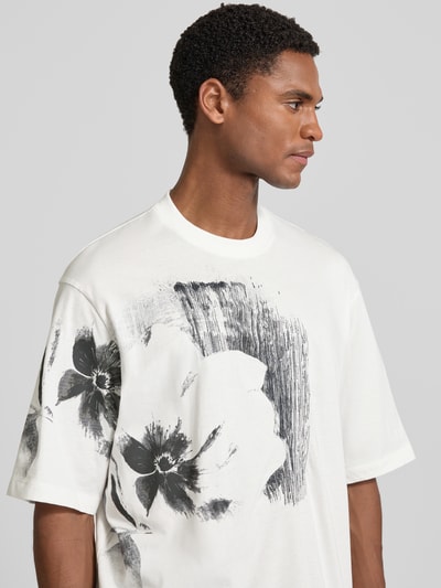 Mannenmodel draagt Comfort Fit T-shirt met motiefprint van Armani Exchange in all-over look, witte en zwarte tinten, ronde hals, ruglengte maat M 76 cm, detailopname bovenlichaam.