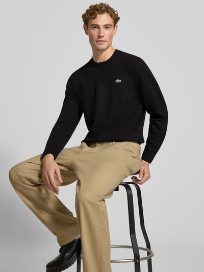 Männermodel trägt schwarzen Regular Fit Pullover aus reiner Wolle von Lacoste, beigefarbene Hose, Ganzkörperansicht von vorne. Herren Pullover von Lacoste