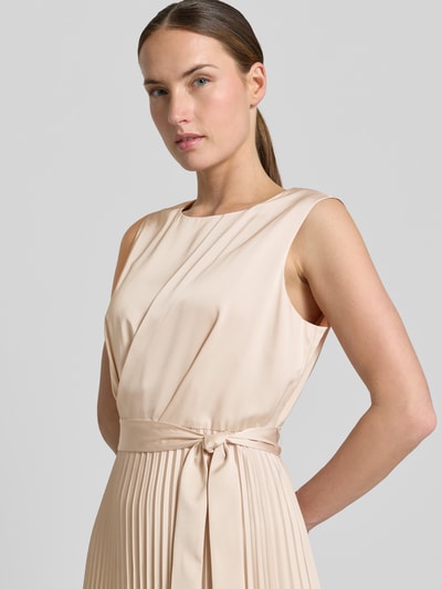 Modelka nosi Midikleid mit Plisseefalten und Bindegürtel von comma, mieszanka materiałów z dodatkiem streczu, linia a, plisy, wiązany pasek, szerokość w rozmiarze 36: 40 cm, długość tyłu w rozmiarze 36: 35 cm, kremowy kolor, gładka faktura, zbliżenie na górną część tułowia, widok z przodu.