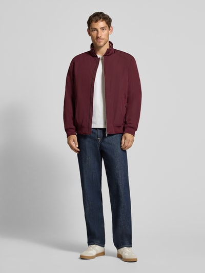 Männermodel trägt Regular Fit Jacke mit Stehkragen von Christian Berg Men in Dunkelrot, kombiniert mit Jeans und Sneakers, Ganzkörperansicht von vorne. Rückenlänge 69 cm, Schulterbreite 49 cm, Brustweite 55 cm, Ärmellänge 67 cm, elastische Abschlüsse, Reißverschluss, Eingrifftaschen.