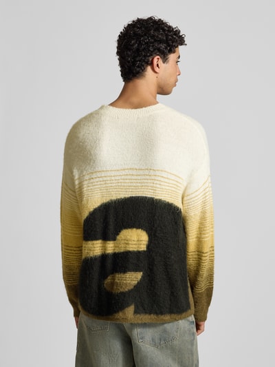 Männermodel trägt Oversized Strickpullover mit gerippten Abschlüssen von ANOTHER COTTON LAB, Material-Mix mit Baumwoll-Anteil, Farbverlauf in Gelb und Beige, Label-Stitching auf Rückseite, Rückenlänge 68 cm, Ärmellänge 57 cm, Ganzkörperansicht Rückansicht.