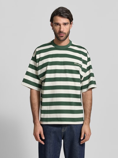 Männermodel trägt Relaxed Fit T-Shirt mit Logo-Stitching Modell 'Beam' von Woodbird aus reiner Baumwolle, grün-weiß gestreift, Ganzkörperansicht von vorne.
