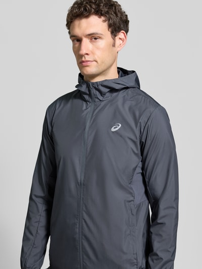 Männermodel trägt dunkelgrauen Windbreaker mit Logo-Print und Kapuze von ASICS, glatte Oberfläche, Seitentaschen, Nahaufnahme Oberkörper.