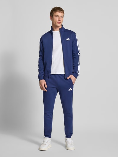 Männermodel trägt blauen Trainingsanzug mit Label-Details von adidas Sportswear, Gerade Passform, Stehkragen, Ganzkörperansicht von vorne.