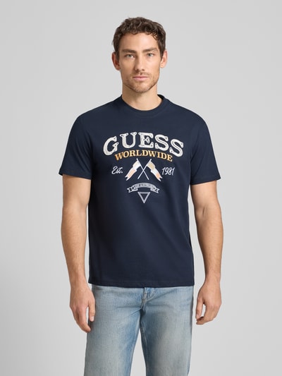 Męski model nosi T-Shirt mit Rundhalsausschnitt und Label-Stitching od Guess z bawełny, krój regular fit, model 'FLAGS AND CORD', ciemnoniebieski, widok całej sylwetki z przodu.
