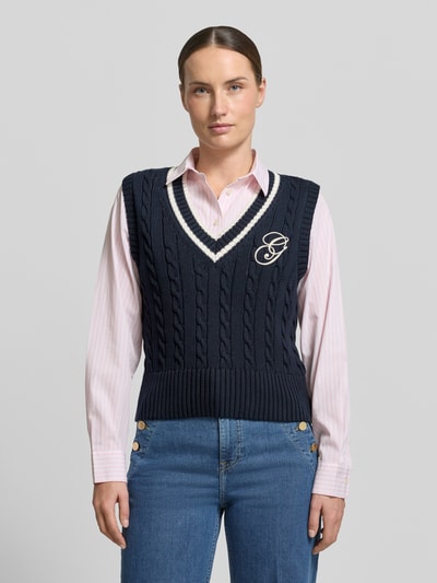 Kobiece model nosi bezrękawnik Pullunder mit V-Ausschnitt und Label-Stitching von Gant z bawełny, krój slim fit, ścieg warkoczowy, wyhaftowane logo, dekolt w serek, długość tyłu w rozmiarze S: 50 cm, widok całej sylwetki z przodu.