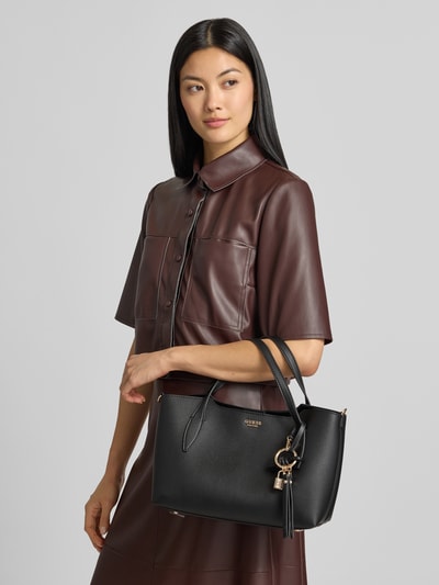Vrouwenmodel draagt zwarte Umhängetasche met Label-Applikation van GUESS met hoofvak, magneetsluiting, draagband en clutch, Ganzkörperansicht van voren.