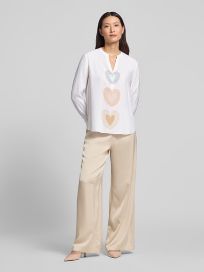 Kobiece modelka nosi Relaxed Fit Tunika-Bluse mit Allover-Motiv-Print von HERZENS, dekolt z lamówką, krój relaxed fit, nadruk z motywem na całej powierzchni, tył wydłużony, z długim rękawem, długość tyłu w rozmiarze 36: 68 cm, długość rękawów w rozmiarze 36: 63 cm, jasne kolory, proste formy, gładka faktura, Ganzkörperansicht von vorne.