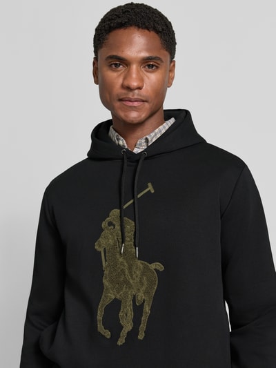 Männermodel trägt Regular Fit Hoodie aus Baumwoll-Mix von Polo Ralph Lauren in Schwarz mit großem Logo-Print auf glatter Oberfläche, Nahaufnahme Oberkörper. Herren Hoodie von Polo Ralph Lauren
