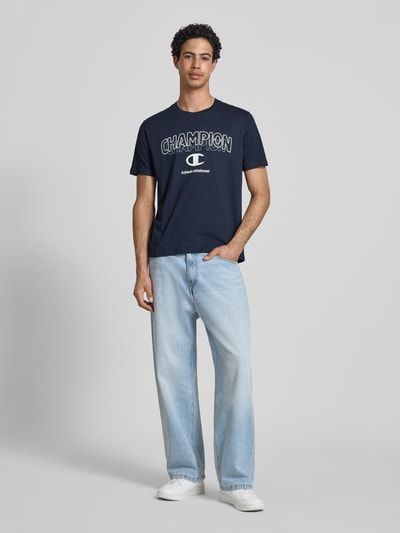 Männermodel trägt T-Shirt mit Label-Print und Rundhalsausschnitt von CHAMPION, Reine Baumwolle, Regular-Fit, dunkelblau, Ganzkörperansicht von vorne.