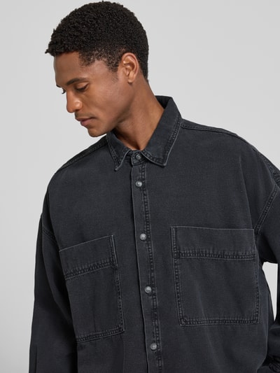 Männermodel trägt Relaxed Fit Jeanshemd mit Kentkragen Modell 'JAZY' von American Vintage aus Reine Baumwolle in dunklem Ton, Nahaufnahme Oberkörper, sichtbare Brusttaschen, Kontext: Rückenlänge 81 cm, Schulterbreite 67 cm, Ärmellänge 58 cm bei Größe M/L.