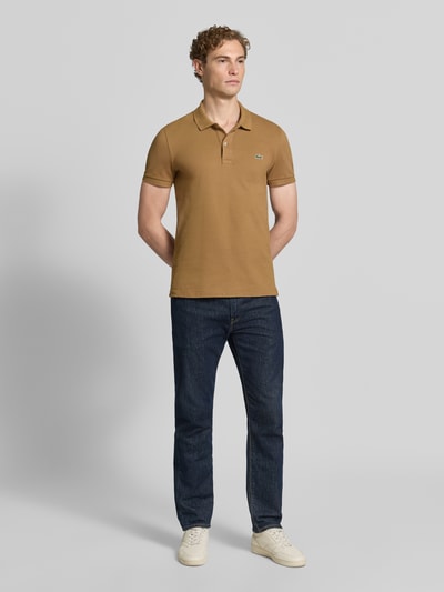 Männermodel trägt Slim Fit Poloshirt aus reiner Baumwolle von Lacoste in Braun und Jeans, Ganzkörperansicht von vorne.