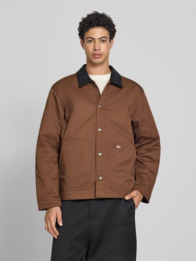 Mannenmodel draagt Jacke mit Umlegekragen und Label-Detail van Dickies in bruin materiaalmix met katoen, regular fit, knoopsluiting, steekzakken, labeldetail; ruglengte maat M 75 cm, schouderbreedte 47 cm, borstwijdte 64 cm, mouwlengte 66 cm. Ganzkörperansicht van voren.
