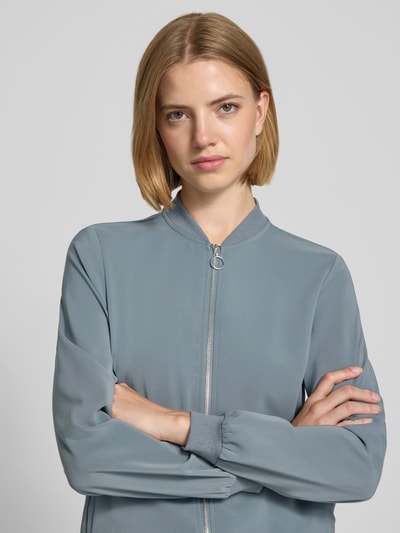 Kobiece model prezentuje Regular Fit Blouson mit Allover-Muster model 'COCO' od VERO MODA w kolorze niebieskoszarym, tekstura gładka, zamek błyskawiczny, wzór na całej powierzchni, damska bomberka; zbliżenie na górną część ciała, widok od przodu.