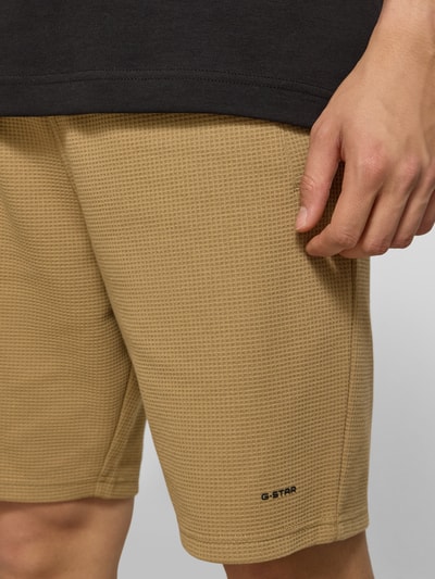 Mannenmodel draagt Straight Fit Shorts met structuurpatroon van G-Star Raw in beige, elastische materiaalmix met katoen, logodetail, elastische band, bandwijdte maat M 44 cm, binnenbeenlengte maat M 20 cm, detailopname dijbeen