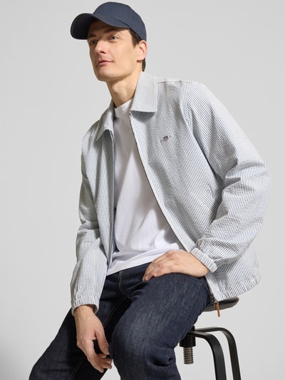 Männermodel trägt Jacke mit Zweiwege-Reißverschluss und Label-Stitching von Gant aus Baumwoll-Elasthan-Mix, Regular-Fit, Ganzkörperansicht von vorne. Rückenlänge 68 cm, Schulterbreite 43 cm, Brustweite 60 cm, Ärmellänge 66 cm, Umlegekragen, Eingrifftaschen, dezentes Streifenmuster.