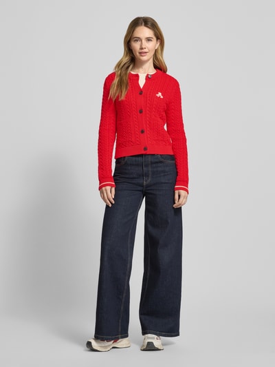 Vrouwenmodel draagt Slim Fit Strickjacke aus reiner Baumwolle Modell 'JULIAANAS CABLE' van Armedangels, effen rood met kabelpatroon, geribde boorden, lange mouwen, ronde hals, opgestikte zakken, vooraanzicht hele lichaam