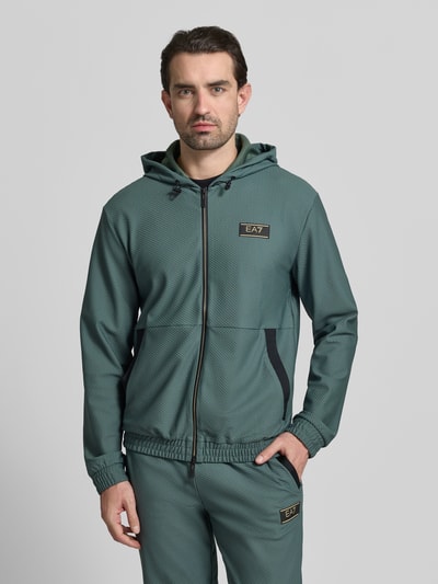 Männermodel trägt Regular Fit Sweatjacke mit Logo-Patch von EA7 Emporio Armani in Grün mit Strukturmuster, Eingrifftaschen, Kapuze, Nahaufnahme Oberkörper.