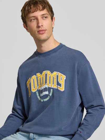 Männermodel trägt Regular Fit Sweatshirt aus reiner Baumwolle von Tommy Jeans in Dunkelblau mit gelbem Print, Nahaufnahme Oberkörper. Herren Sweatshirt von Tommy Jeans.