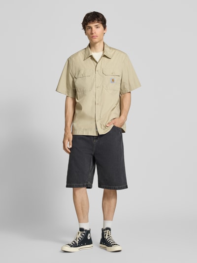 Männermodel trägt Relaxed Fit Freizeithemd mit Kentkragen und Label-Detail von Carhartt Work In Progress im Material-Mix mit Baumwoll-Anteil, Farbe Beige, große Brusttaschen, kombiniert mit dunkler Shorts, Rückenlänge 75 cm, Schulterbreite 51 cm, Ärmellänge 28 cm, Ganzkörperansicht von vorne.